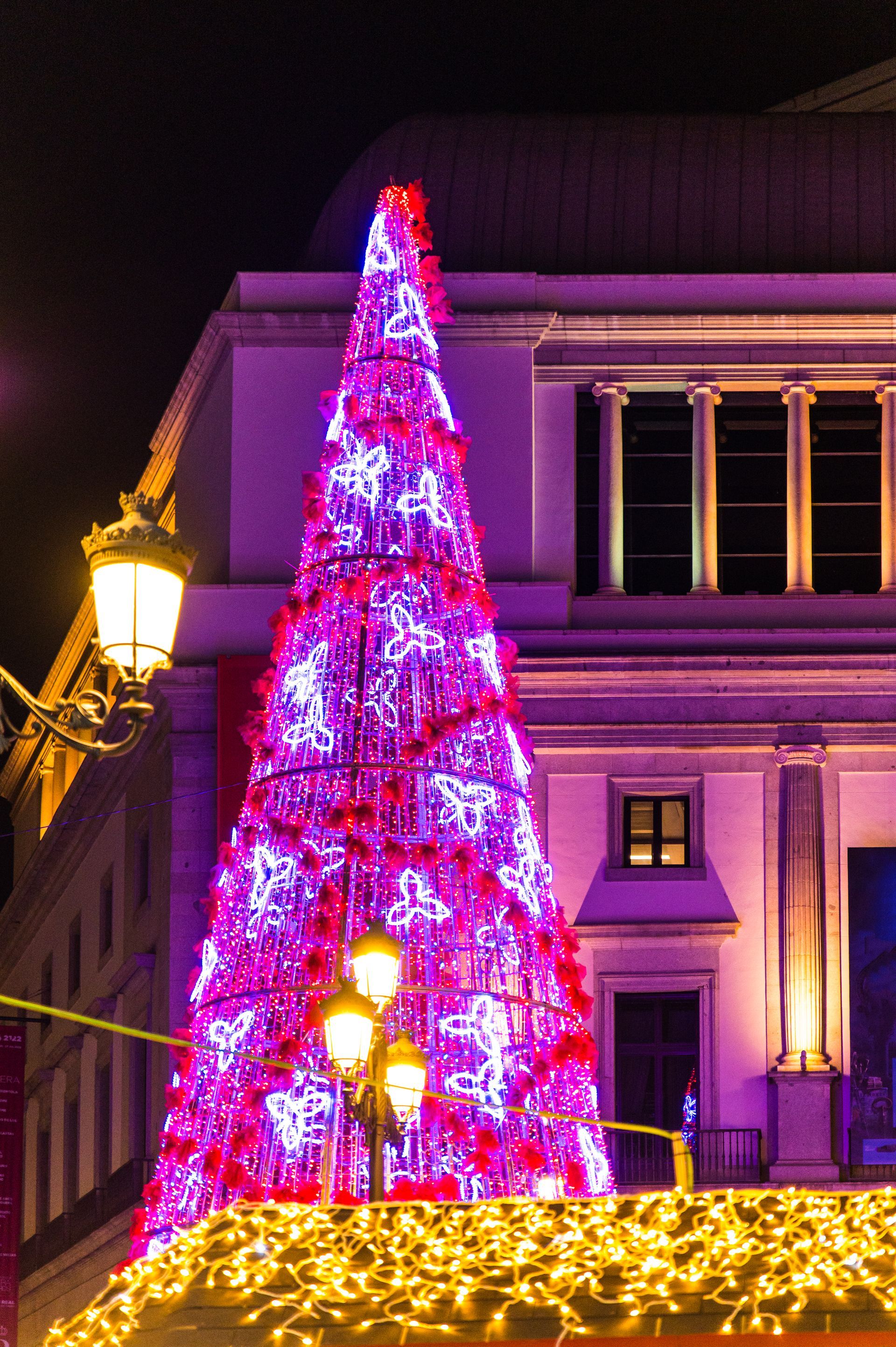 A brightly lit pink Christmas tree in front of a building at night, with golden lights along the base.