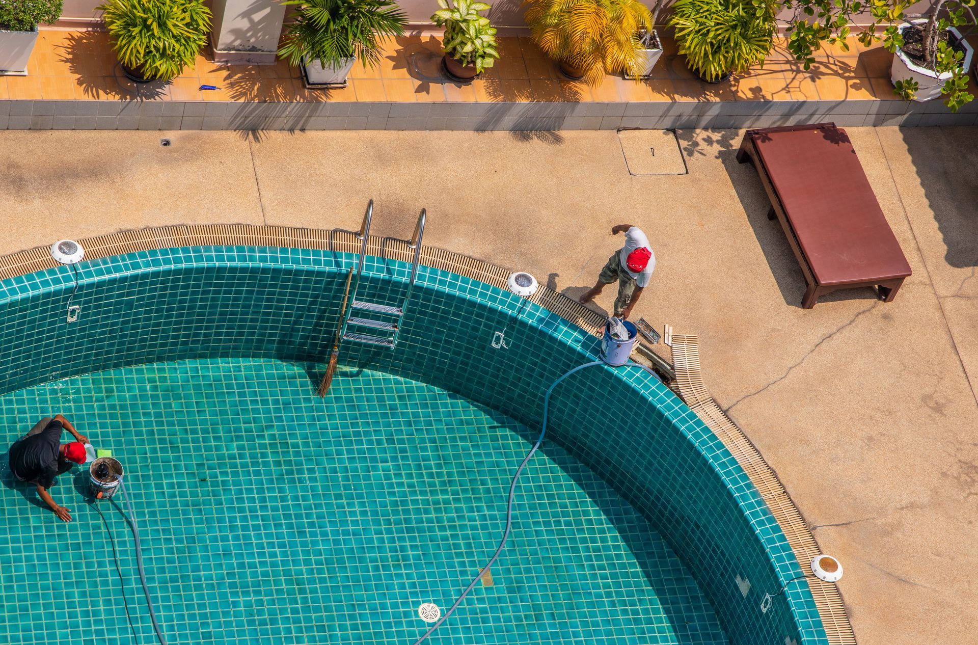 Two workers cleaning a turquoise swimming pool beside a sandy deck and lounge chair.