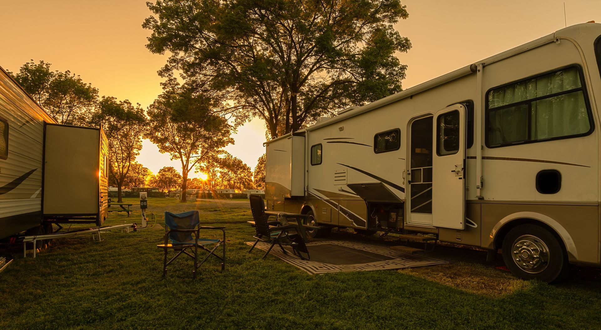 RV campsite at sunset, with two recreational vehicles, chairs, and a table, under a warm golden sky.