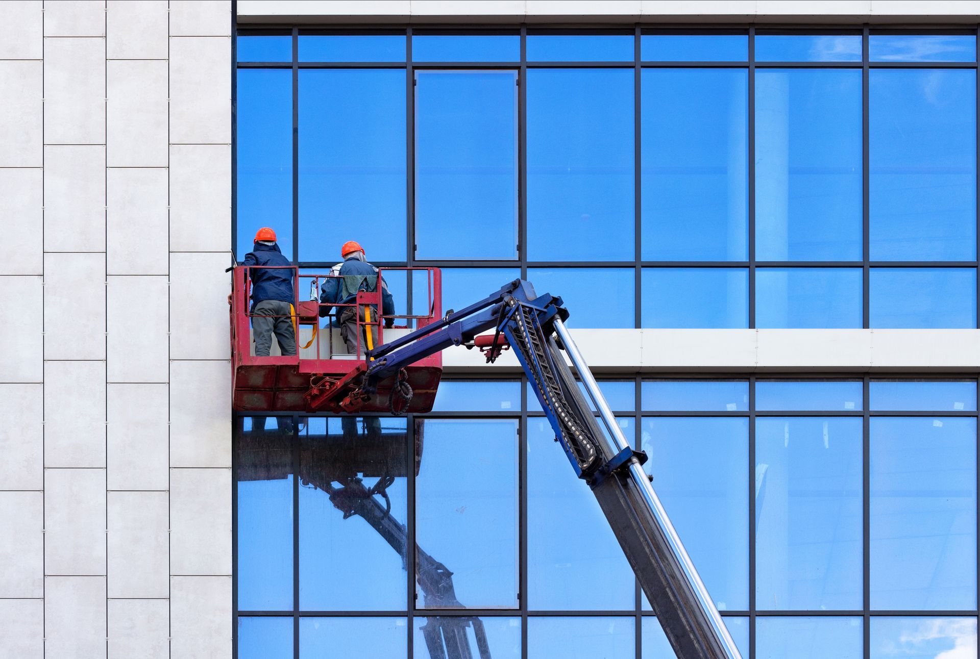 Workers on a red lift beside a blue glass building, cleaning windows with a boom arm.