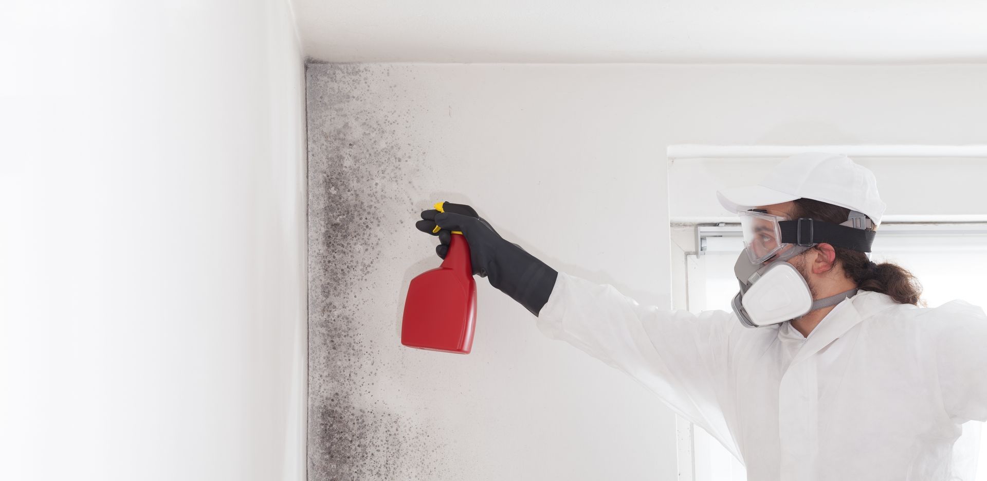 Person in protective gear spraying mold on a white wall.
