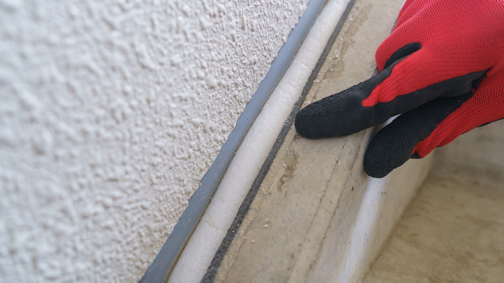 Gloved hand touching a white wall edge near a metal strip on an indoor floor
