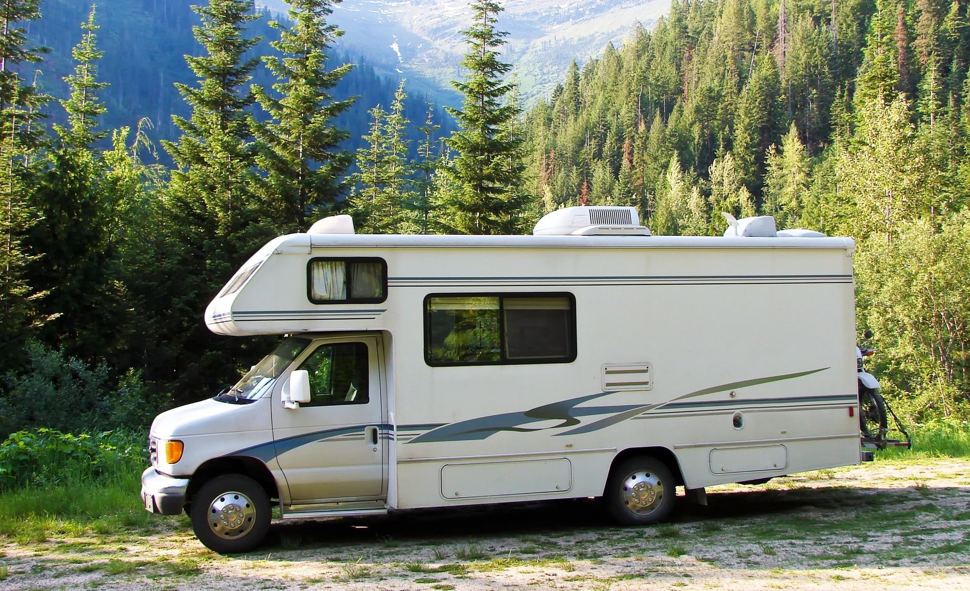 White RV parked in a grassy area, surrounded by pine trees and mountains in the background.