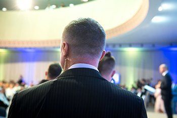 Man in a dark suit seen from behind at a conference, with blurred attendees and colorful stage lighting.