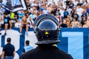 Helmeted security officer faces a cheering crowd at a sports stadium, with blue-and-white banners behind.