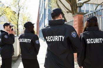Four security guards in black jackets walking outside along a city sidewalk.