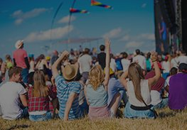 Crowd sitting on grass at an outdoor festival, with colorful flags and a stage in the background