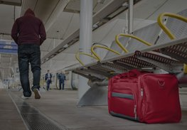 Red suitcase on a train platform beside benches, with a person walking away in the background.