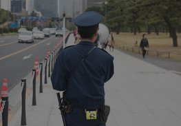 Police officer standing on a city sidewalk near a road, facing away from the camera.
