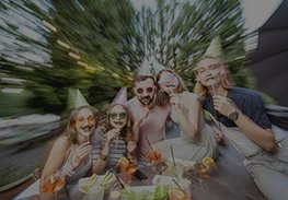 Friends laughing around a table outdoors, holding drinks, with a zoom-blur effect around them.