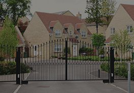 Gated entrance to a residential neighborhood with brick houses and trees behind black metal fencing.