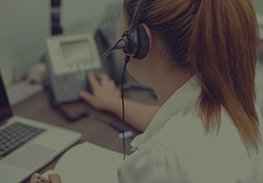 Call center agent wearing a headset at a desk, typing beside a desk phone and laptop