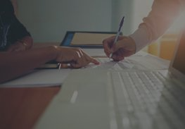 Hands writing on paper beside a laptop in a bright office setting