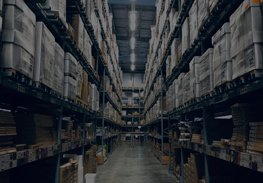 A tall warehouse aisle with stacked pallets and shelves on both sides, lit by overhead lights.