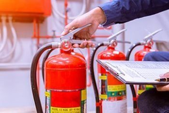 Hands checking a red fire extinguisher during inspection, with clipboards and other extinguishers nearby