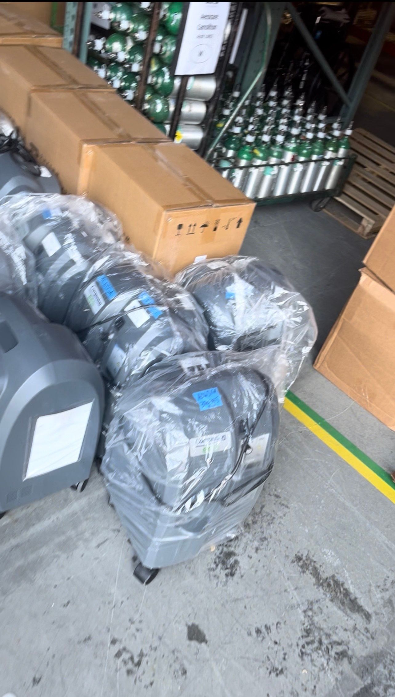 Several wrapped gray machines on floor, cardboard boxes, green cylinders in the background.