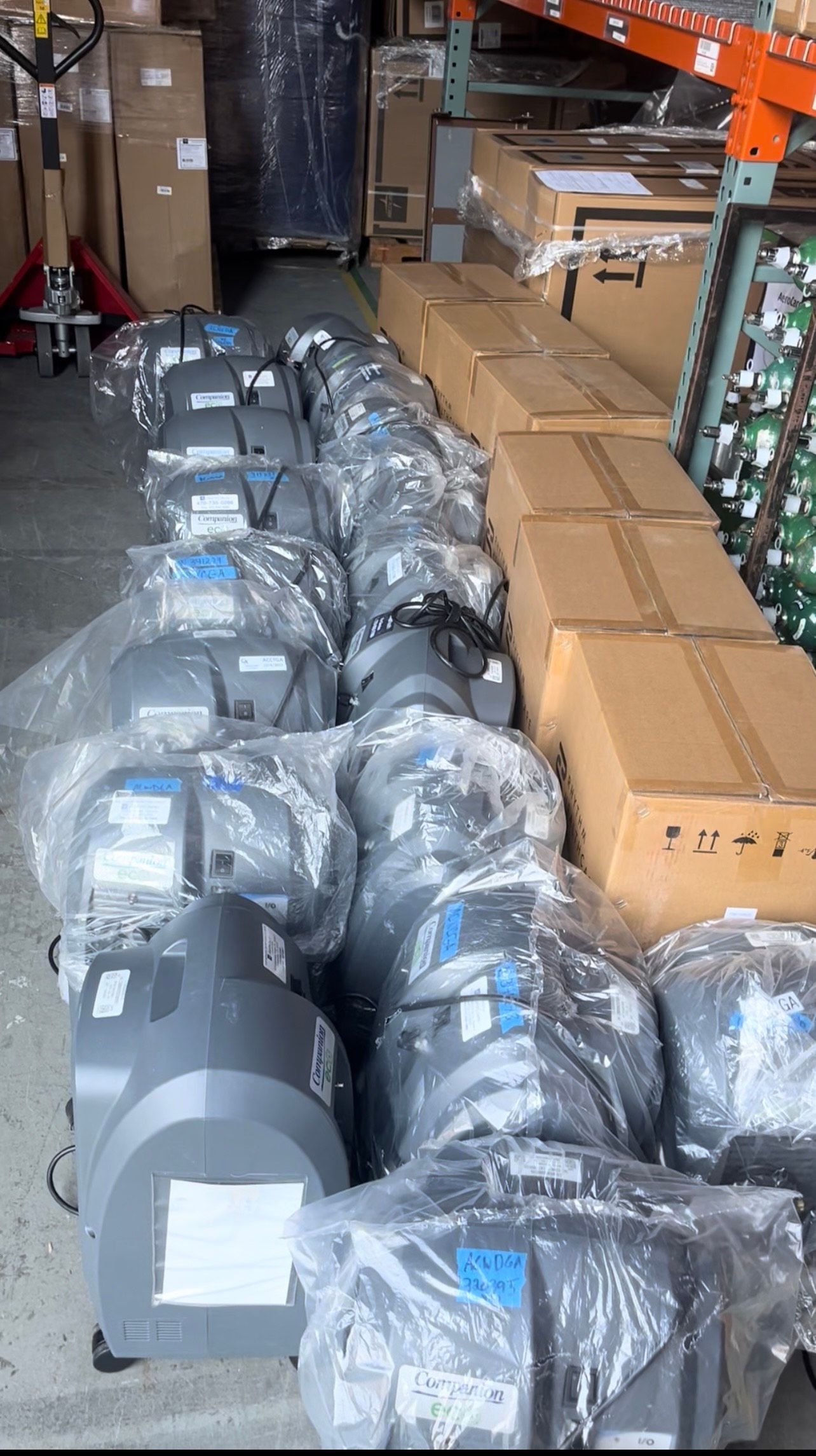 Rows of plastic-wrapped, gray machines and cardboard boxes are stacked in a warehouse.