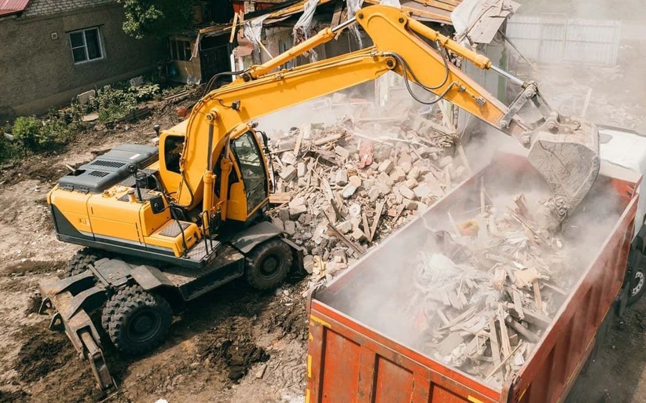 a yellow excavator is loading rubble into a dumpster .