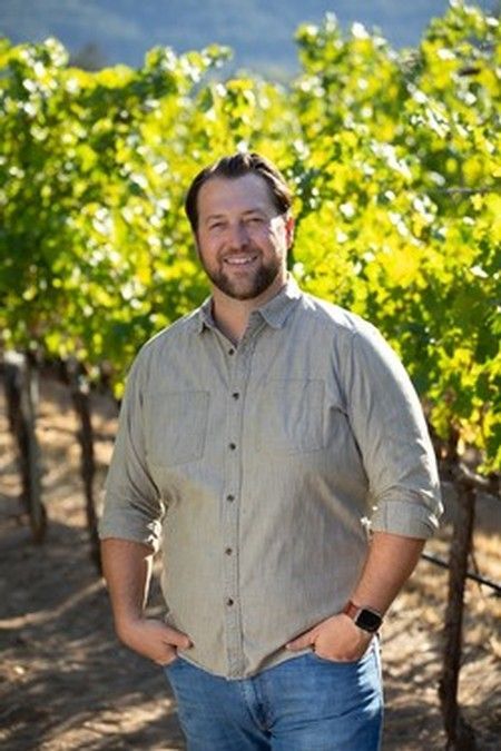 Man in gray shirt and jeans standing in a vineyard, hands in pockets, smiling.