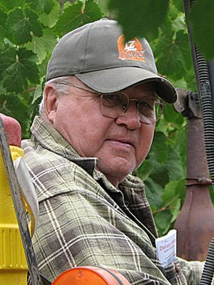 Man in olive cap and glasses, sitting outdoors in front of leafy vines.