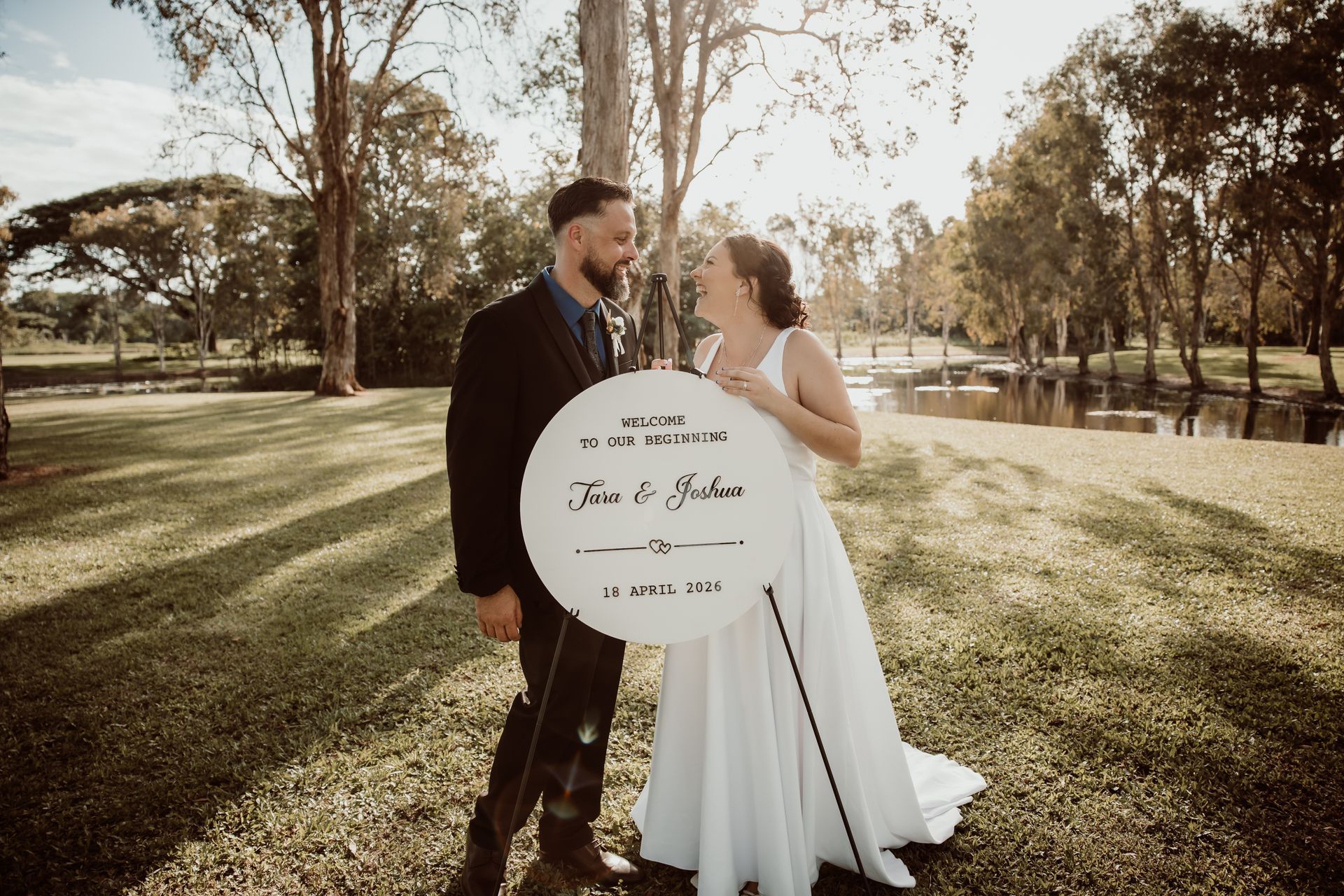 Bride and groom in a sunlit park, smiling beside a round sign in front of a pond