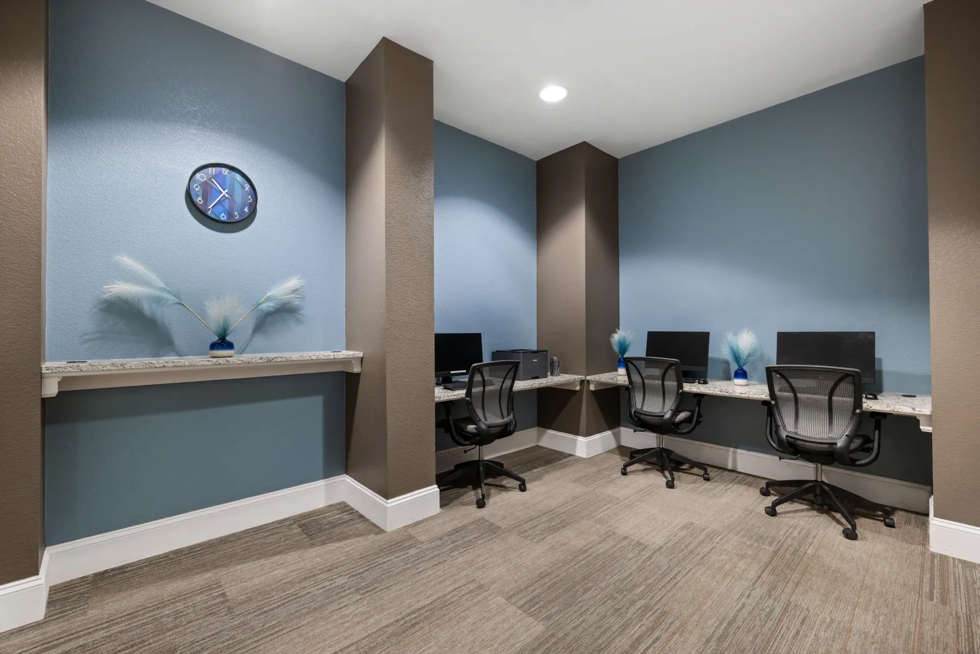 Communal workspace with three desks, chairs, and computer monitors along blue and brown walls at The Parc at Greenwood Village.