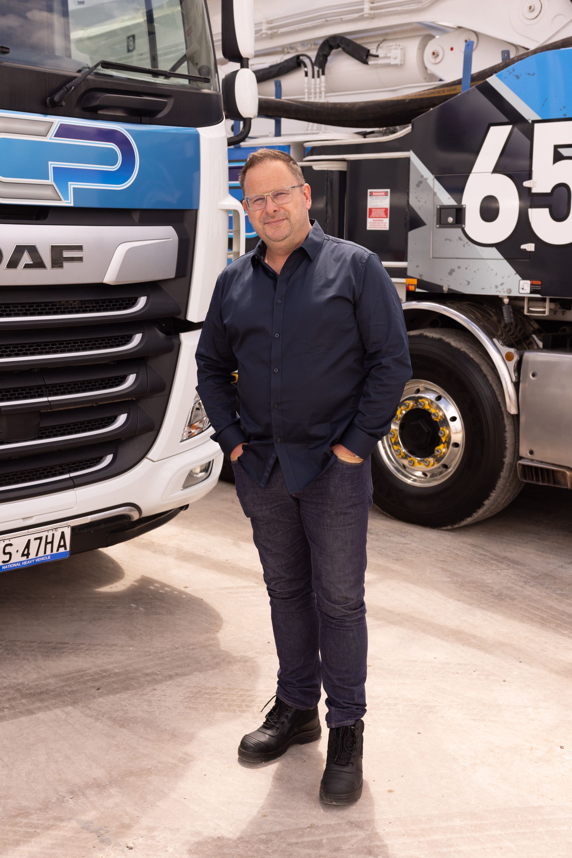 Man standing with hands in pockets in front of a truck, wearing a blue shirt and jeans.