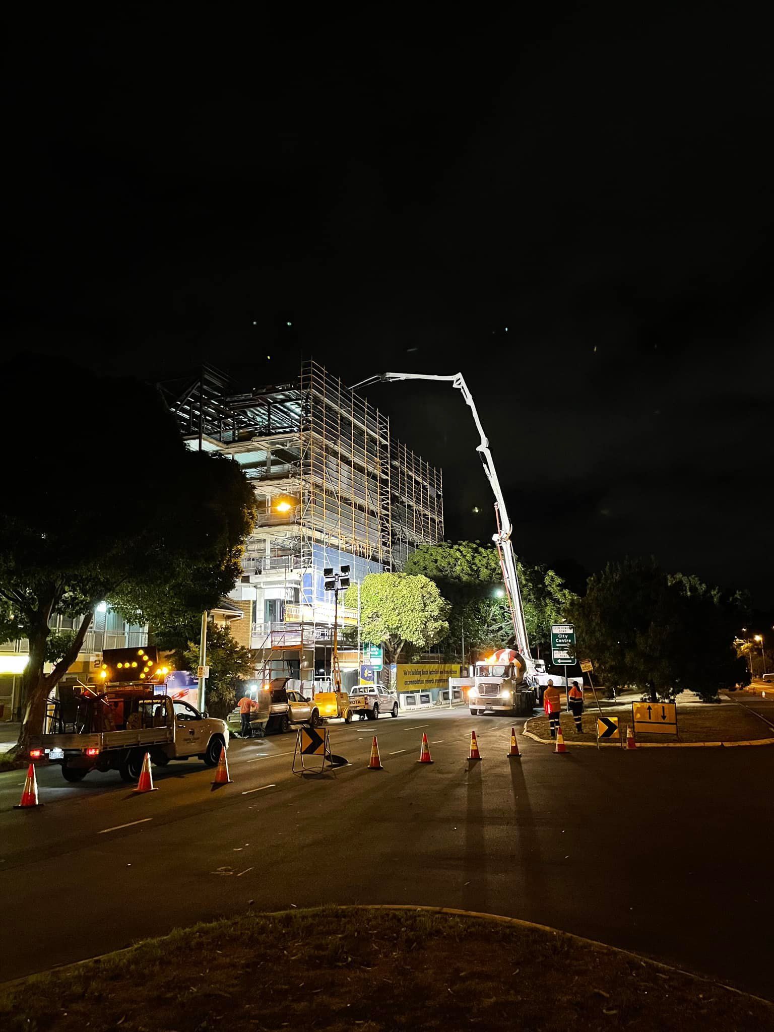 Nighttime construction site with a concrete pump truck reaching a building frame.