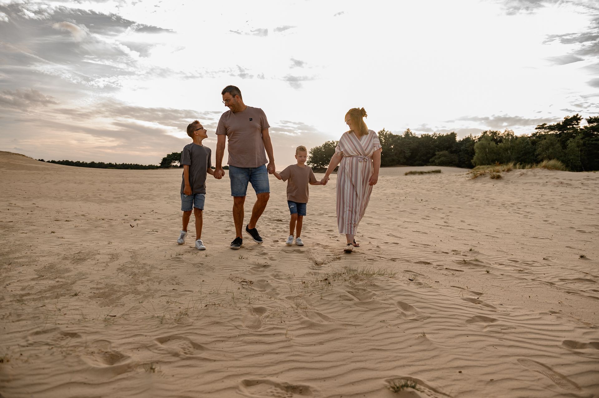 Een gezin van vier loopt hand in hand over een zandstrand. Op de achtergrond is een bewolkte hemel te zien.