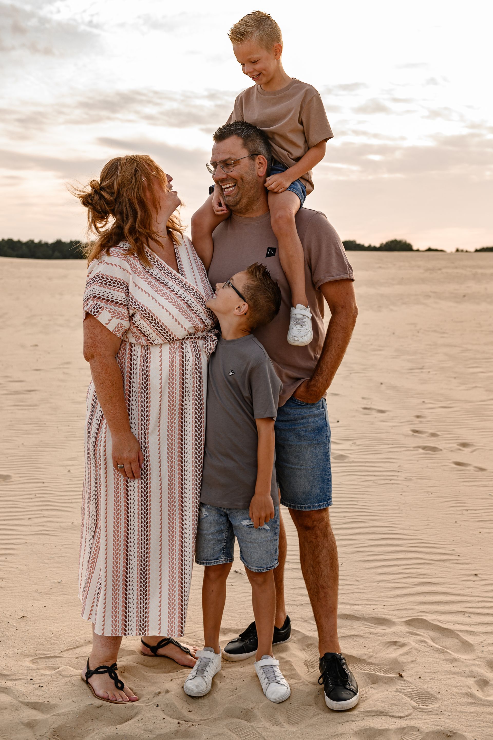Een gezin van vier in vrijetijdskleding op een zandstrand. De zoon zit lachend op de schouders van zijn vader. Zonsondergang.
