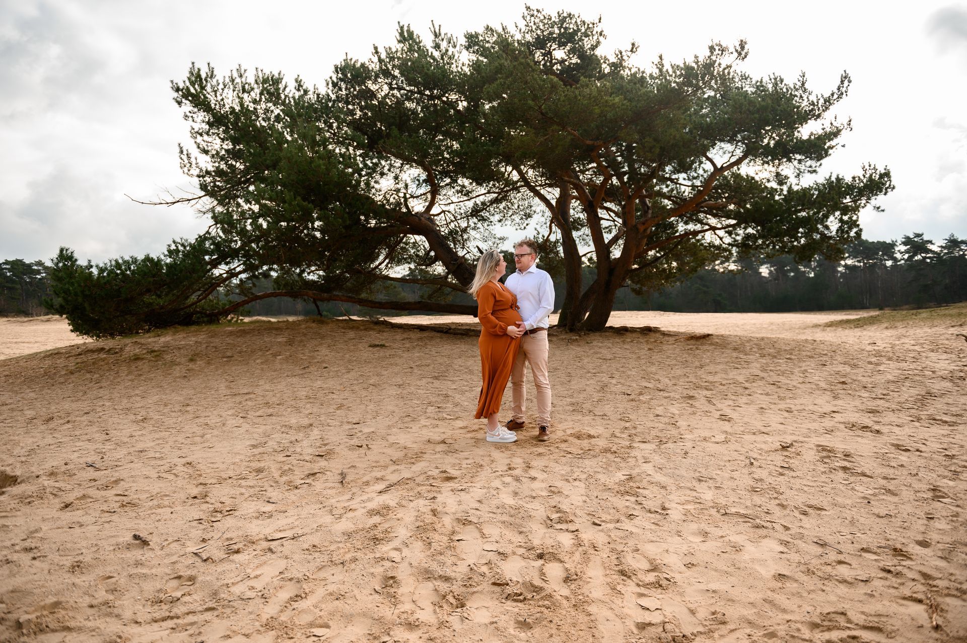 Een stel staat onder een boom in een zanderig landschap; de man draagt ​​een beige broek en een wit shirt, de vrouw een jumpsuit.