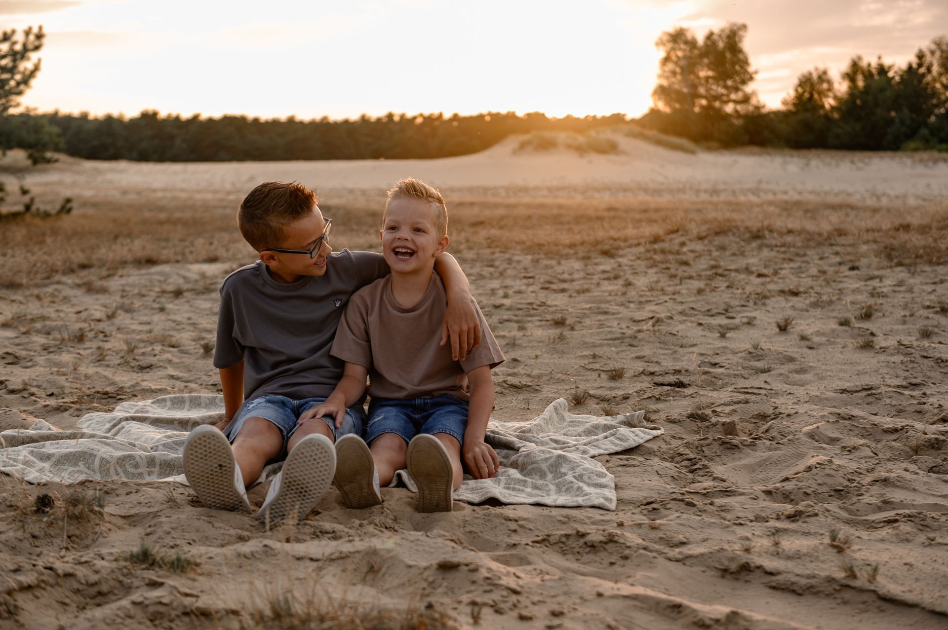 Twee jongens zitten op een deken in het zand, lachend en elkaar omhelzend, met een zonsondergang op de achtergrond.