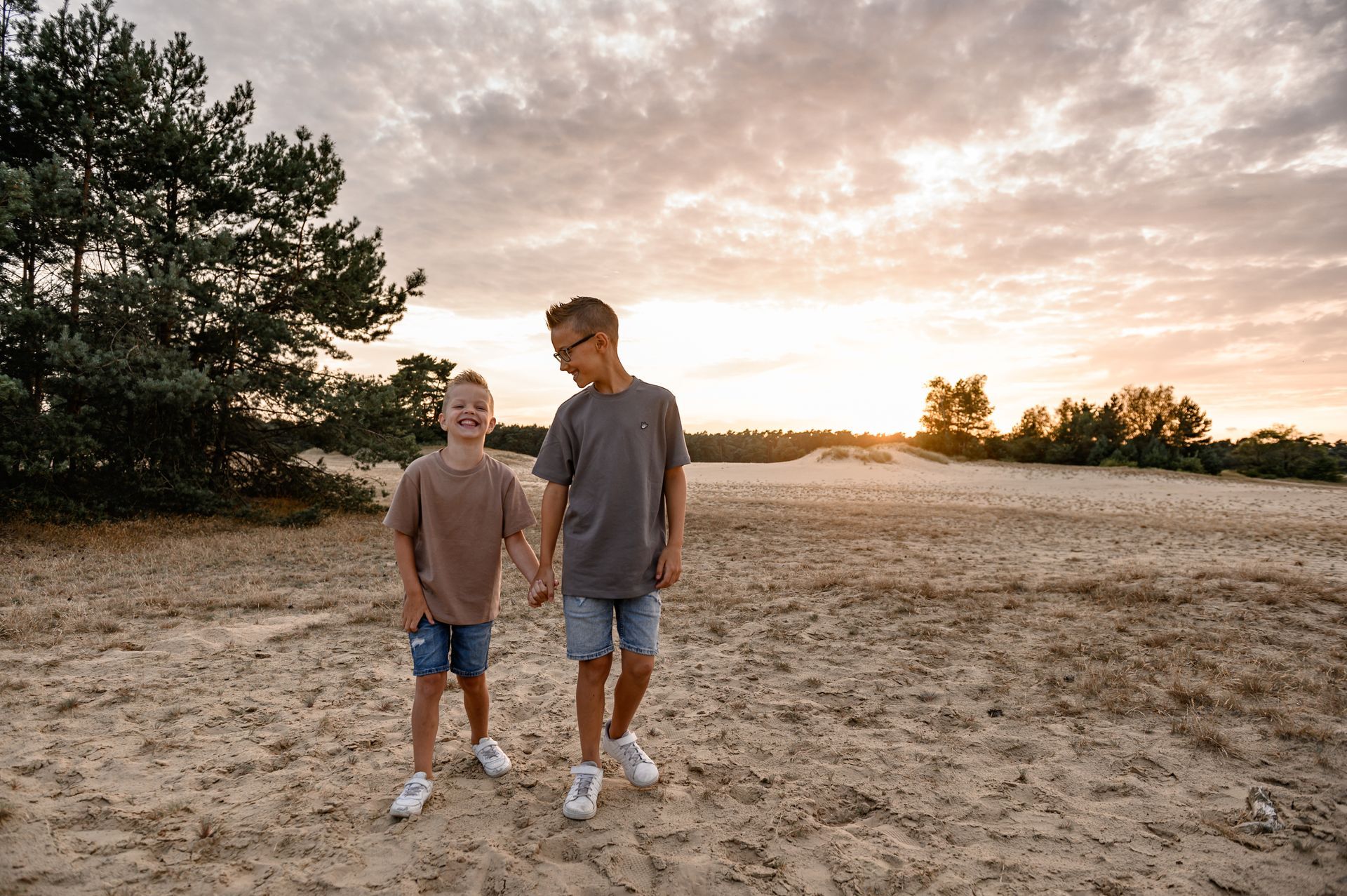 Twee kinderen lopen hand in hand over een zanderig landschap bij zonsondergang.