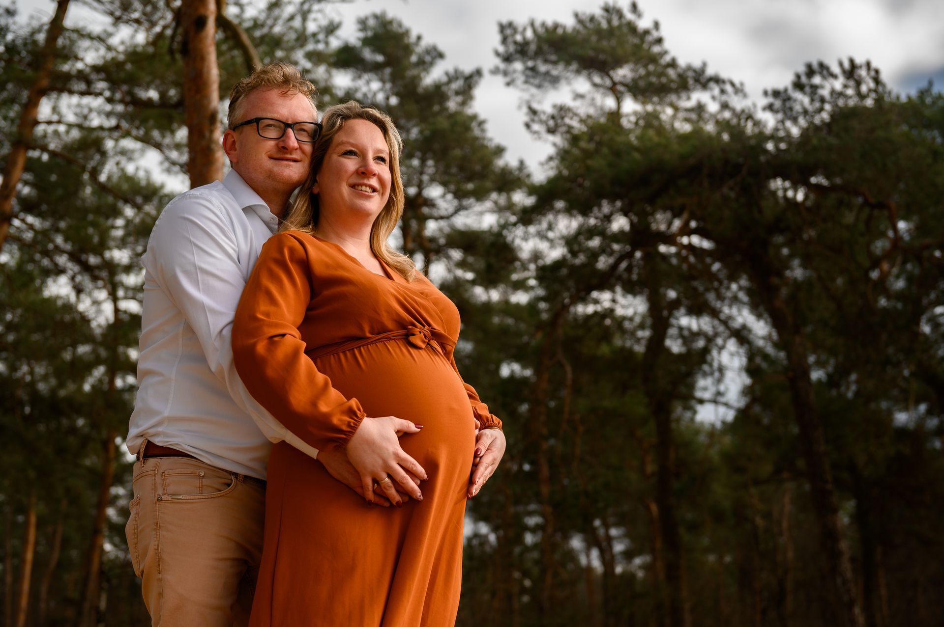 Een zwangere vrouw en haar partner poseren buiten in een bos; de vrouw draagt ​​een oranje jurk, de man een wit shirt en beiden glimlachen.