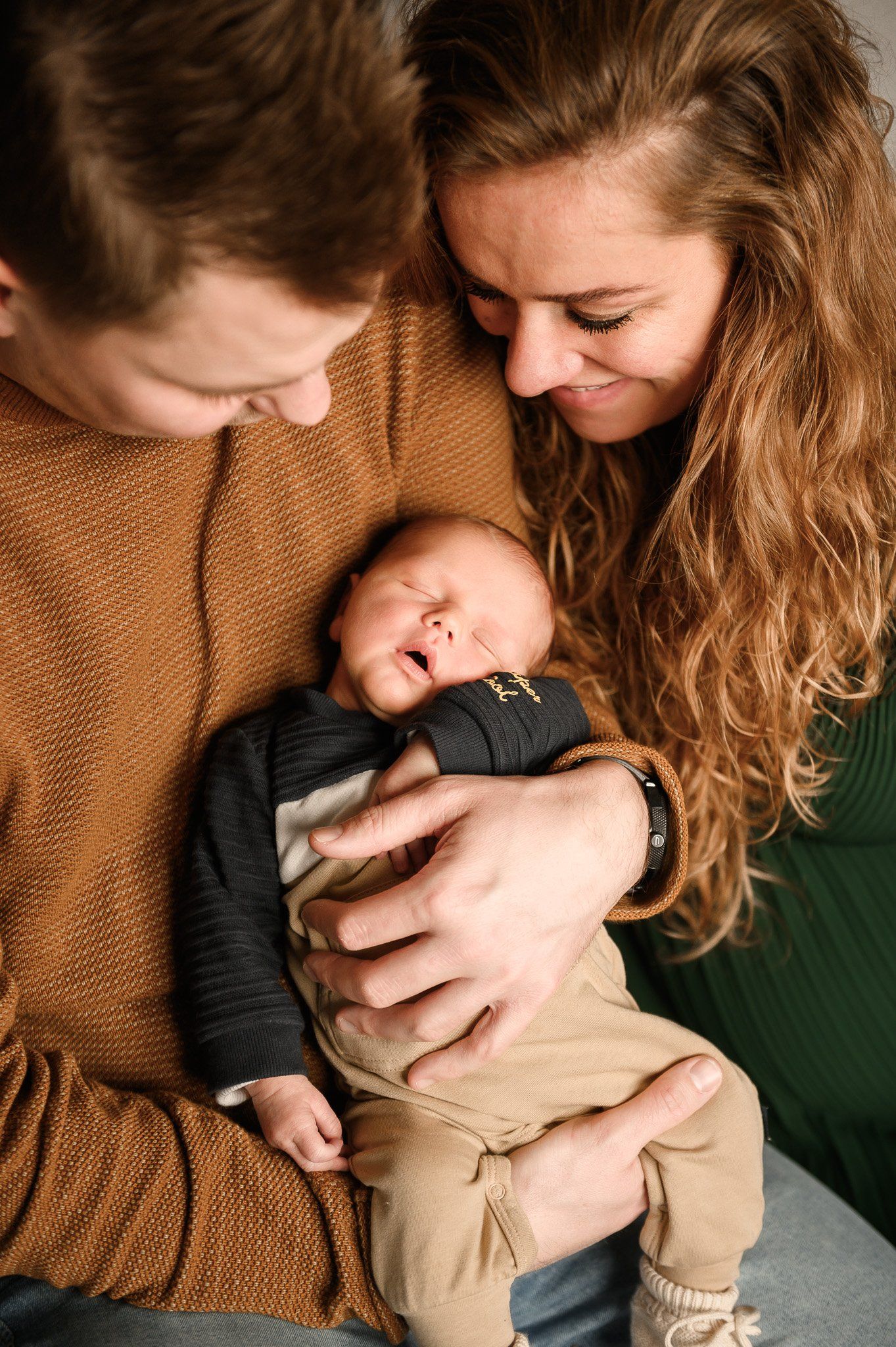 Ouders kijken vol bewondering naar hun pasgeboren baby, die ze in hun armen houden. De baby draagt ​​bruin-groene kleding.
