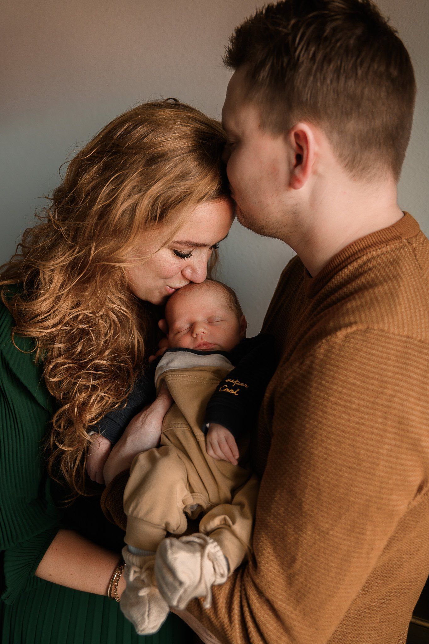 Ouders kussen het hoofdje van een pasgeboren baby. De vrouw met krullend haar draagt ​​groen. De man draagt ​​bruin.