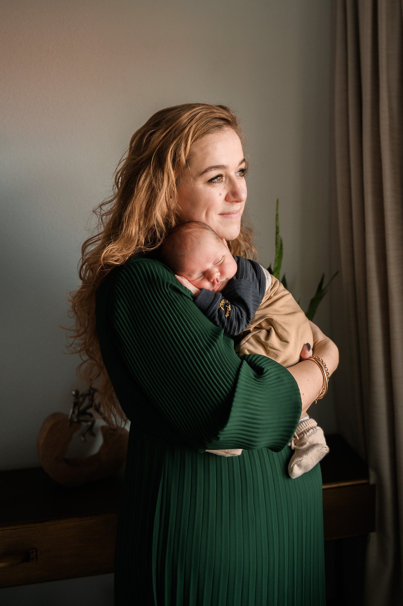 Een vrouw met krullend haar houdt een slapende baby vast bij een raam; ze draagt ​​een groene jurk.