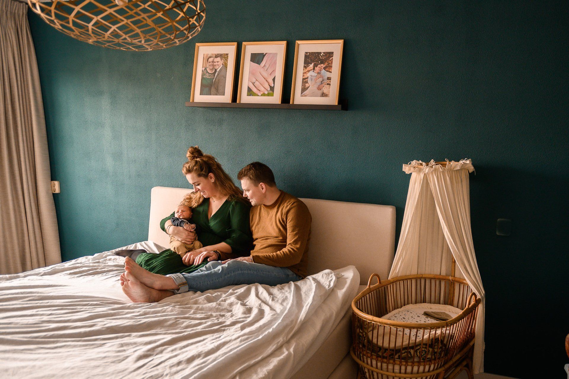 Een gezin van drie personen op een bed: een kind en twee volwassenen. Een turquoise muur, kunst en een wieg in een moderne slaapkamer.