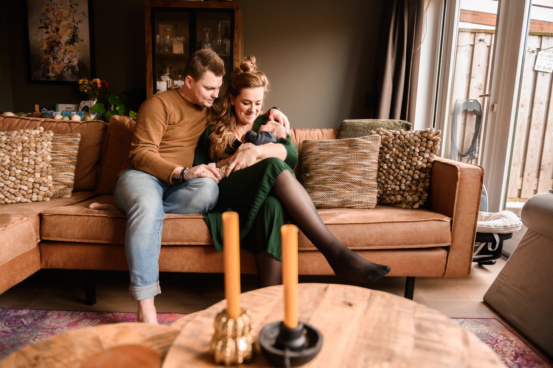 Een stel zit op een bruine bank in de woonkamer met een baby op de arm. Donkergroene muur, houten meubels, kaarsen op een tafel.
