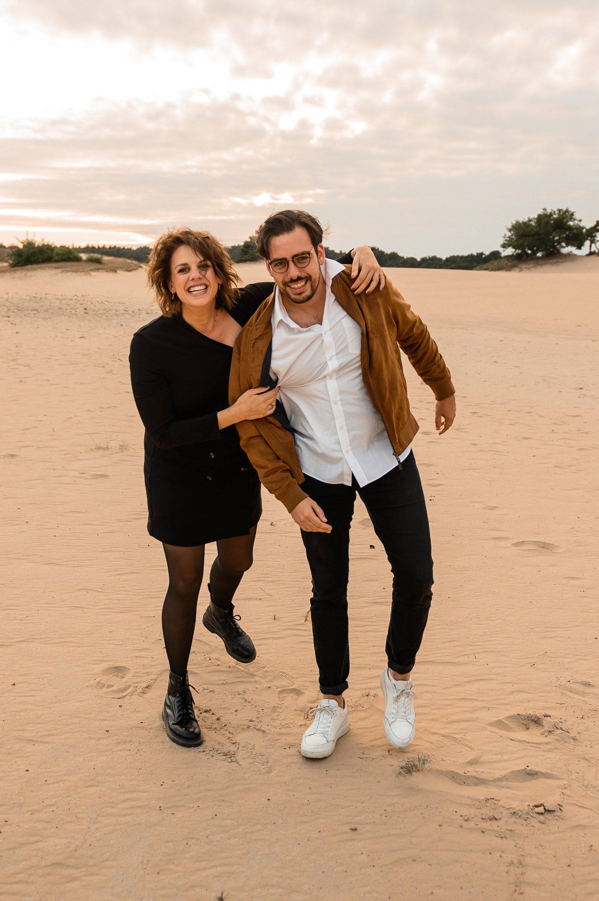 Een stel poseert speels op een zanderig terrein; de man draagt een bruine jas, de vrouw een zwarte jurk.