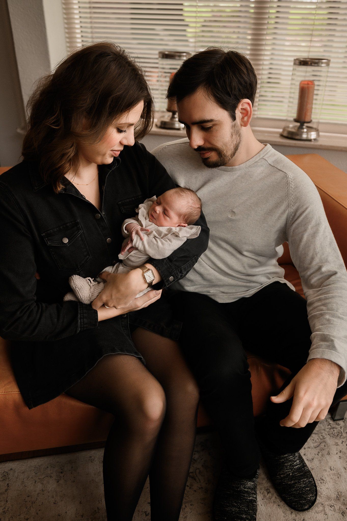 Ouders zitten samen en kijken neer op een pasgeboren baby die ze vasthouden. Ze bevinden zich in een woonkamer.