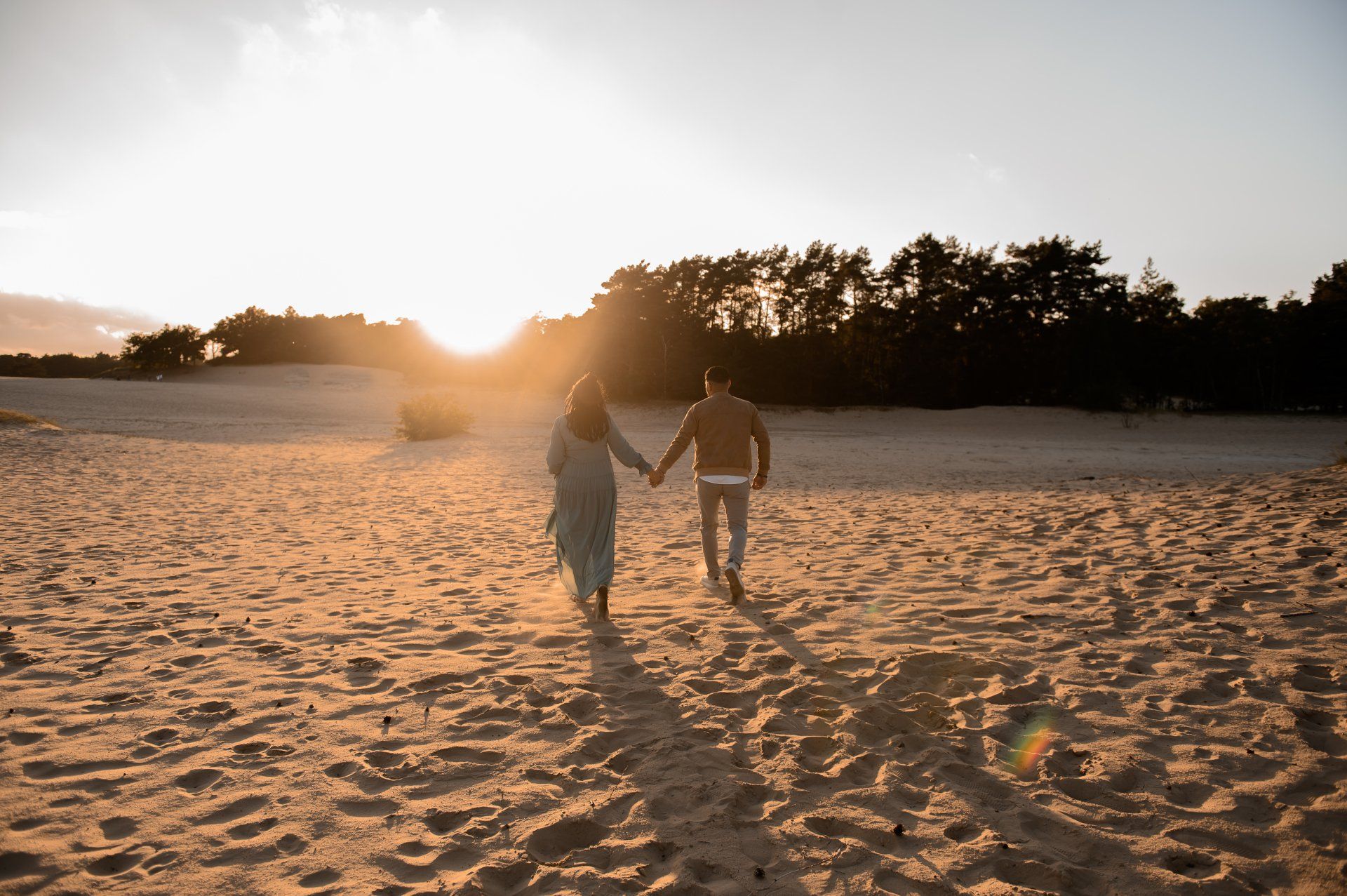 Een stel loopt hand in hand over het zand richting de zonsondergang. Silhouetten tegen een gouden licht.
