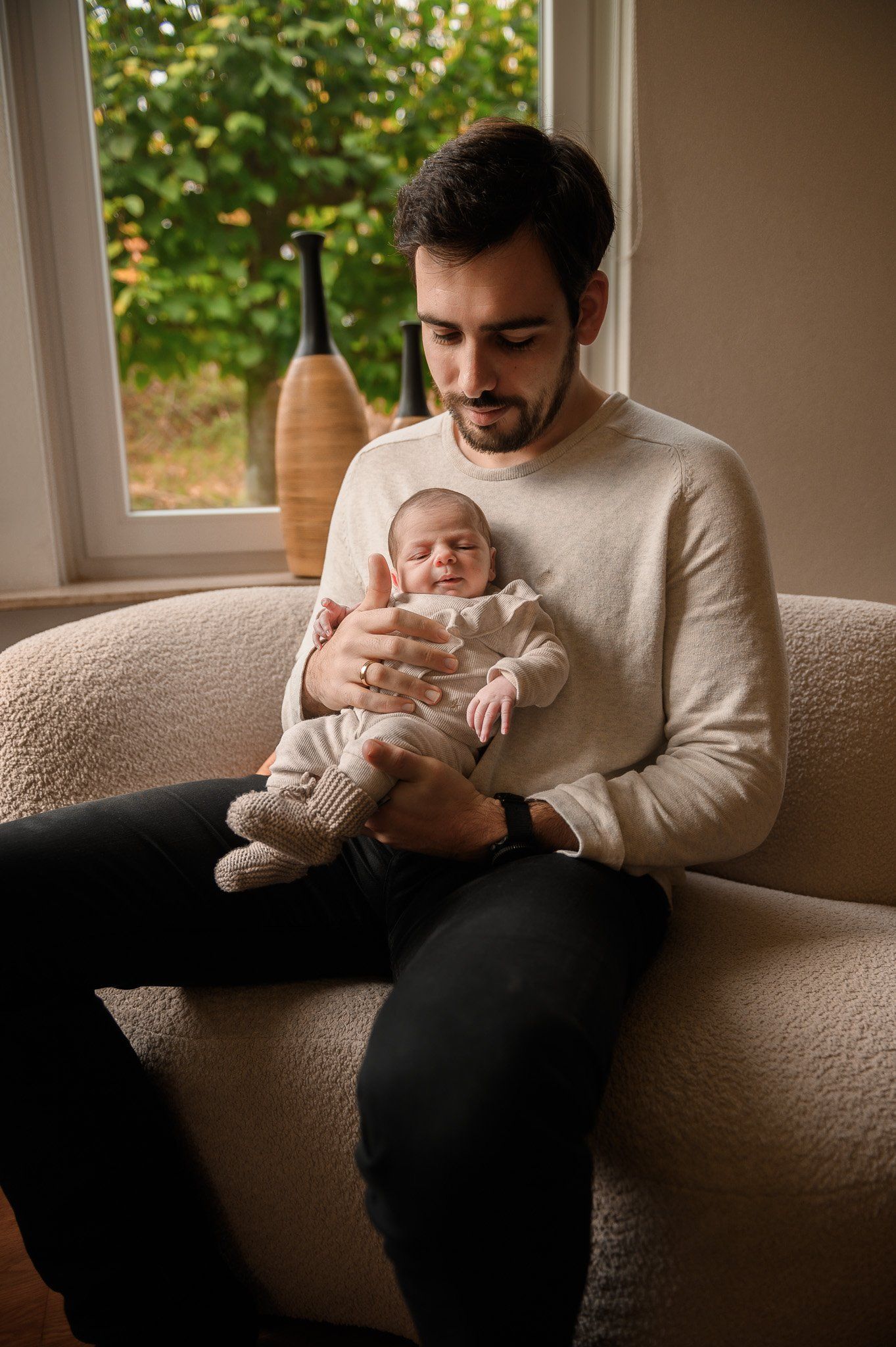 Een man houdt een pasgeboren baby vast op een bank. De man kijkt naar beneden; de baby slaapt. Op de achtergrond is een raam te zien.