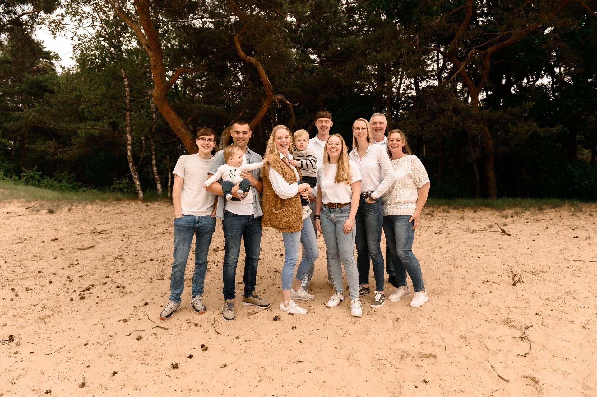 Een gezin van elf personen poseert op een zandstrand onder de bomen; ze zijn allemaal casual gekleed in jeans en lichtgekleurde shirts.