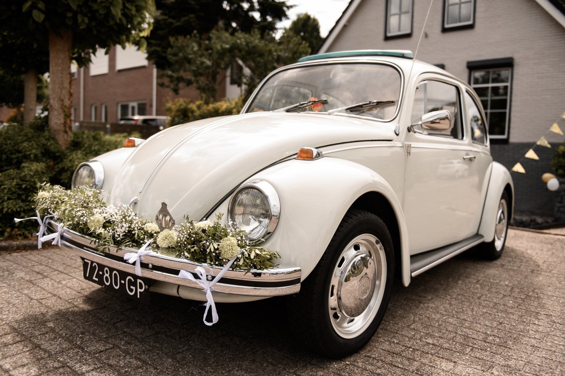 Een witte Volkswagen Kever, versierd met bloemen en linten, staat geparkeerd op een geplaveide oprit.