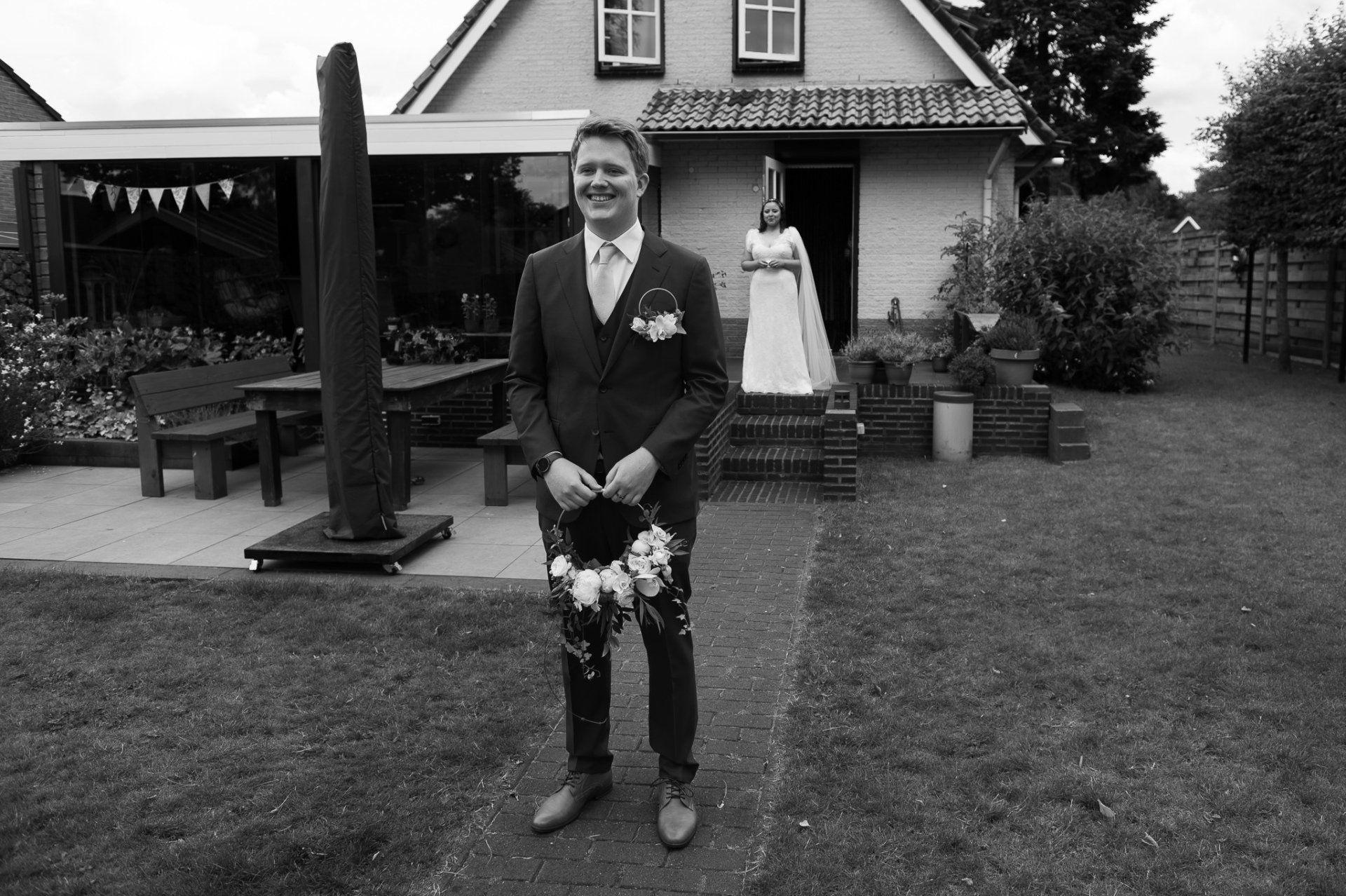 Bruidegom glimlacht, met bloemen in zijn hand, bruid in de deuropening. Trouwfoto buiten een huis met tuin en terras. Zwart-wit.
