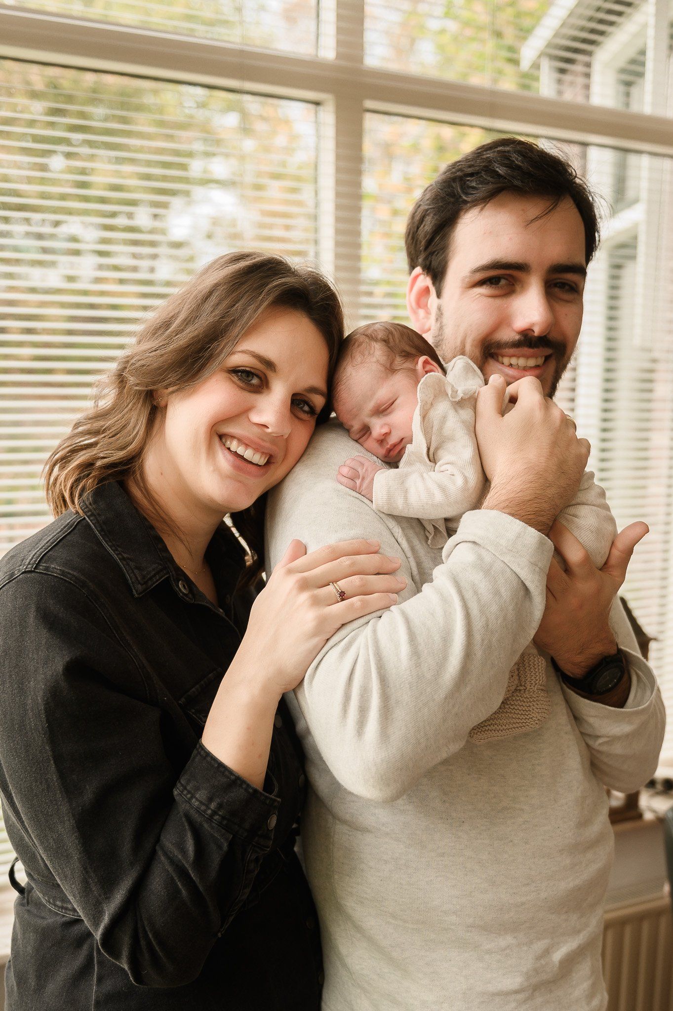 Ouders met een pasgeboren baby in hun armen, glimlachend bij het raam.
