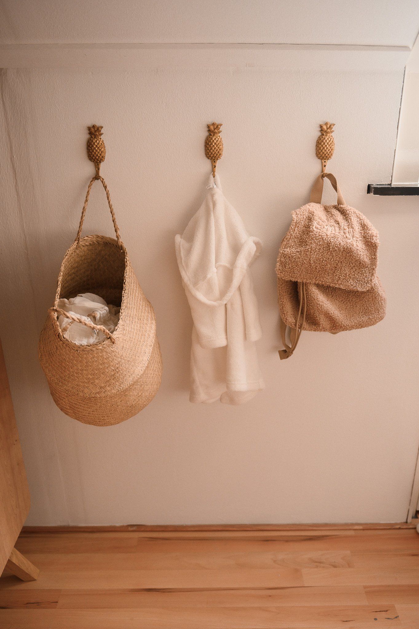 Aan een witte muur hangen drie ananasvormige haken waaraan een gevlochten mand, een witte badjas en een rugzak hangen; de vloer is van hout.