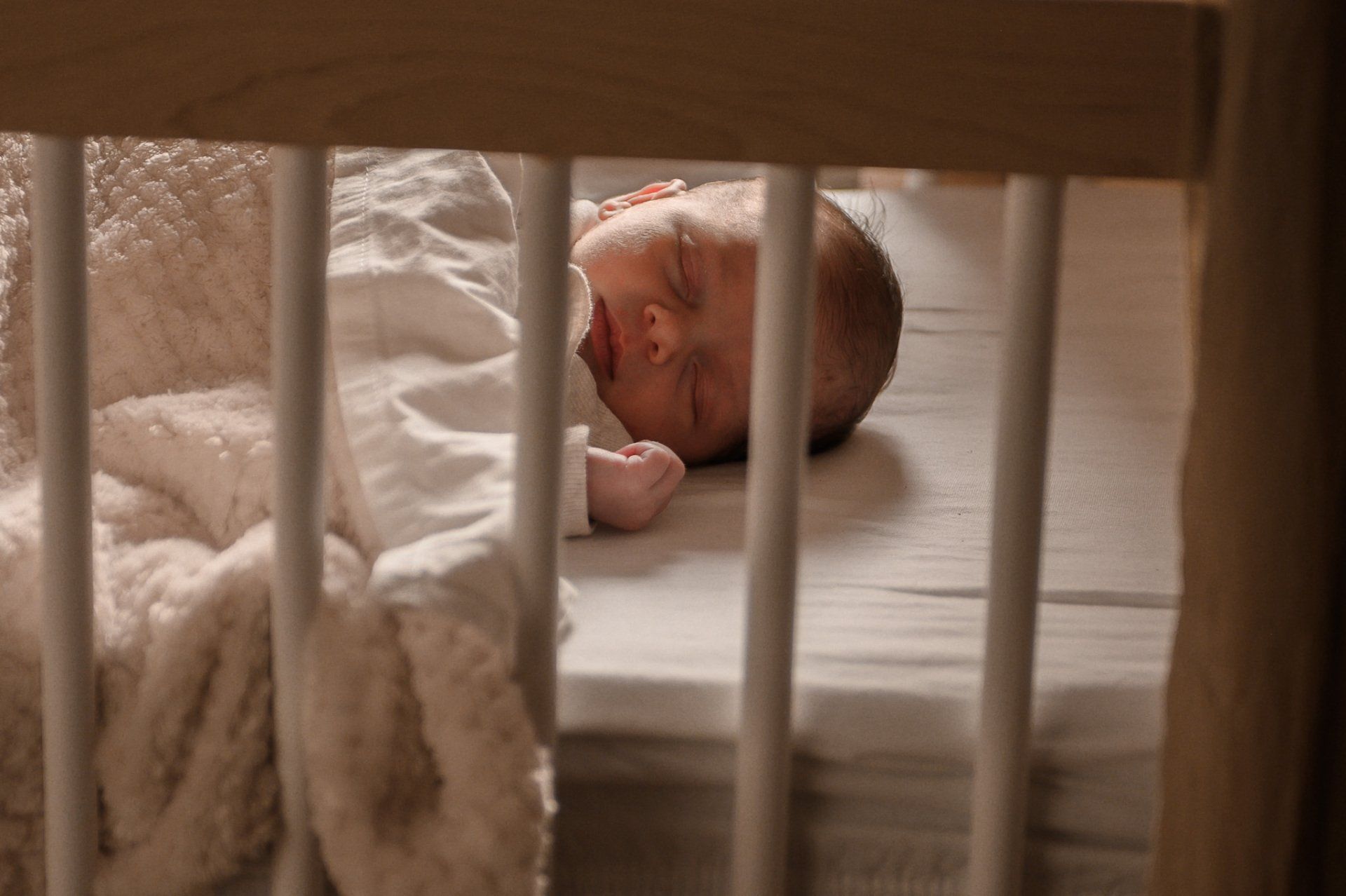 Een baby slaapt in een wiegje met een witte deken, gezien door de houten spijlen.