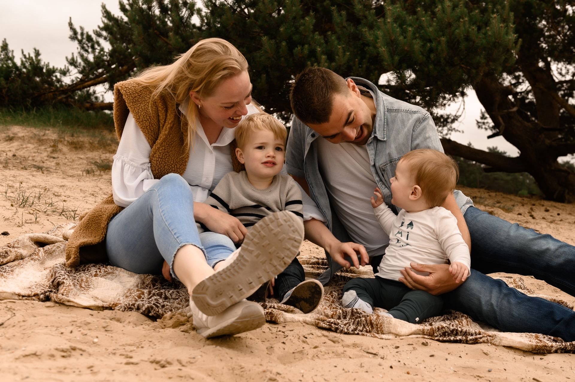 Een gezin van vier zit lachend op het strand. Moeder in een witte blouse en spijkerbroek; vader in een spijkerbroek; twee jonge kinderen.